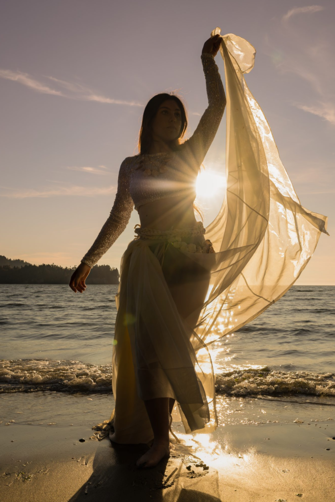 woman in a flowing dress stands on the ocean beach as the sun sets behind her creating a sun star on her side